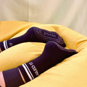 Black Posše grip socks with “Nailed It” cuff and black soles, worn while lying on yellow couch