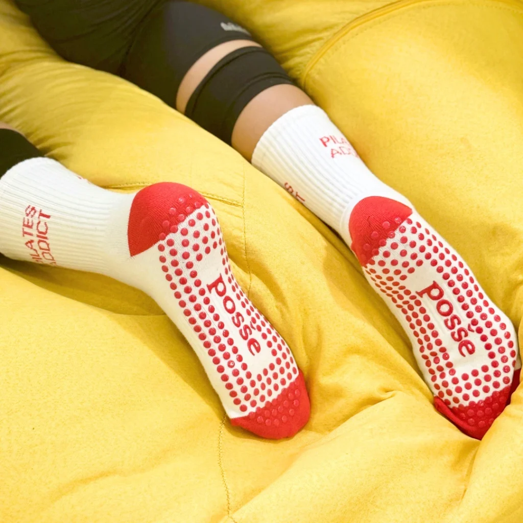 Posše Pilates Addict grip socks in white with red soles and bold red text, worn on yellow couch